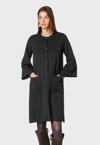 Abito nero al ginocchio realizzato in un morbido tessuto, con scollatura rotonda, maniche a campana lunghe, chiusura con bottoni frontali e due ampie tasche.