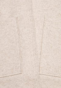 Tessuto di lana beige chiaro con una consistenza morbida, dotato di due tasche frontali e cuciture in vista, che creano un design pulito e semplice.
