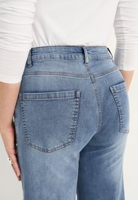 Ljusblå denimjeans med medelhög midja, som har två bakfickor och synlig sömnad. Tyget har en slät och mjuk textur.