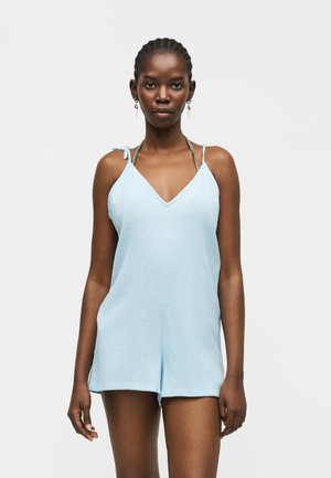 Mujer con cabello trenzado, vestida con un mono sin mangas azul claro con tirantes de lazo, de pie contra un fondo blanco liso.