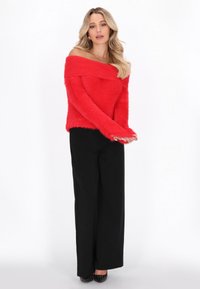 Roter, schulterfreier, kuscheliger Pullover kombiniert mit schwarzen, weit geschnittenen Hosen, die einen entspannten Schnitt und strukturierten Stoff aufweisen.