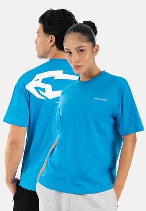 Deux personnes portant des t-shirts bleu vif, l'une face à l'avant avec un petit texte blanc sur la poitrine, l'autre montrant un grand graphisme blanc dans le dos.