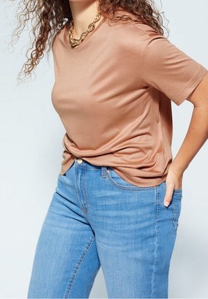Vrouw met krullend haar draagt een beige T-shirt met korte mouwen, een gouden ketting en blauwe spijkerbroek met één hand in haar zak.