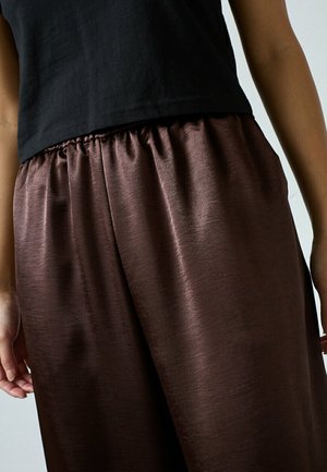 Person iført løstsiddende, skinnende brune satinbukser med elastik i taljen og en sort kort top, stående med armene afslappet langs siderne.