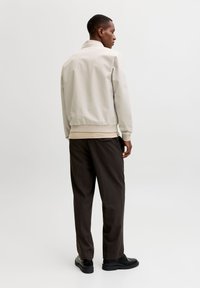 Μπεζ φωτεινό μπουφάν μπέιζερ με ριμπ τελείωμα, φορεμένο πάνω από μπεζ πουκάμισο, σε συνδυασμό με σκούρο καφέ παντελόνι και μαύρα παπούτσια.