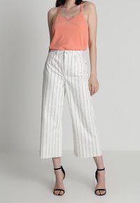 T-shirt corail avec des accents en dentelle, pantalon taille haute blanc avec des rayures verticales noires, et sandales à talons noires.