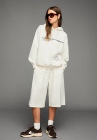 Vrouw in een oversized witte hoodie met de tekst "Art Gallery Launch", bijpassende witte shorts, bruine sneakers, witte sokken en een zonnebril.