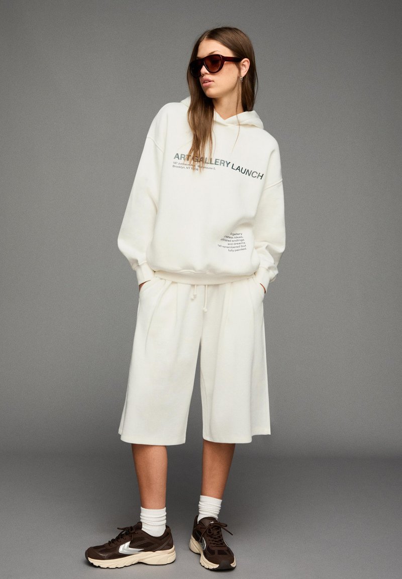 Vrouw in een oversized witte hoodie met de tekst "Art Gallery Launch", bijpassende witte shorts, bruine sneakers, witte sokken en een zonnebril.