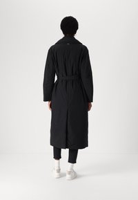 Black long coat with a belted waist, wide collar, and quilted texture. Features a back vent and is styled with dark jeans and white sneakers.