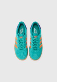 Mėlynos dangaus spalvos zomšos sportbačiai su oranžinėmis juostelėmis, violetiniais akcentais ir guminiais padais. Turi raištelių uždėjimo dizainą ir tekstūruotas vidines padas.