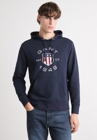 Sudadera azul marino hecha de una mezcla de algodón, con un logo central que incluye un escudo, estrellas y detalles numéricos. Tiene un bolsillo tipo canguro y capucha ajustable.
