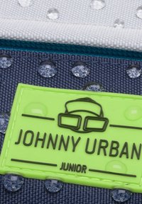 Zielony gumowy metek z napisem "JOHNNY URBAN JUNIOR" na teksturowanej ciemnoniebieskiej tkaninie z widocznymi kroplami wody i białymi akcentami.