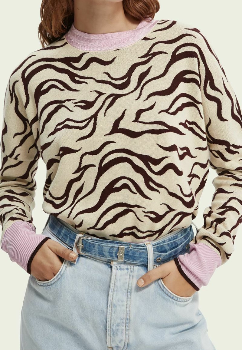Femme portant un pull imprimé zèbre beige et brun foncé avec des poignets et un col roses, rentré dans un jean bleu clair taille haute avec une ceinture en denim.