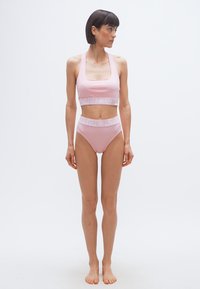 Γυναίκα που στέκεται ξυπόλητη, φοράει ασορτί ανοιχτό ροζ αθλητικό σουτιέν και ψηλόμεσα σλιπ με λευκές σχεδιαστικές λωρίδες, κοιτάζοντας προς το πλάι.