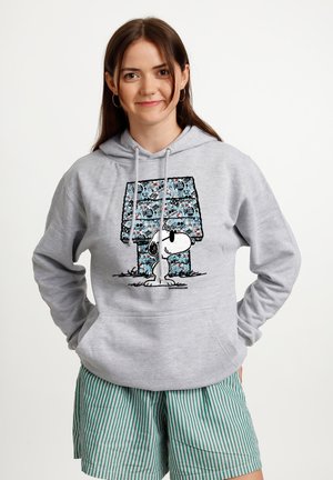 Młoda kobieta uśmiecha się, ma długie brązowe włosy i nosi szarą bluzę z kapturem z psem Snoopy oraz niebieską, wzorzystą budę dla psa, w połączeniu z zielonymi szortami w paski.