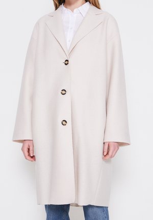 Vrouw draagt een lichtbeige oversized jas met drie grote schildpadknopen over een wit shirt en een blauwe spijkerbroek.