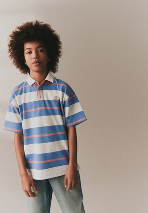 Teenager mit lockigem Haar, der ein blau-weiß-pink gestreiftes Polohemd und hellblaue Jeans trägt, steht vor einem schlichten beigen Hintergrund.