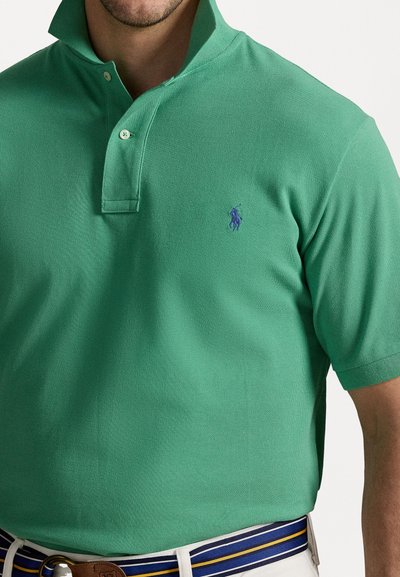 Grønn polo-shirt laget av teksturert stoff med krage, med en liten lilla brodert logo på venstre bryst.