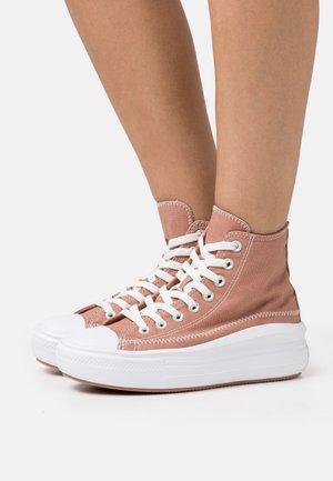 Personne portant des baskets montantes en toile rose poussiéreux avec des semelles épaisses en caoutchouc blanc et des lacets blancs, debout sur une surface blanche.