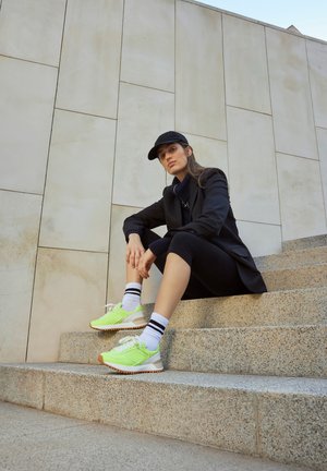 Zapatillas deportivas verde claro con cordones blancos y suelas marrones, combinadas con calcetines blancos con rayas negras. Modelo sentado en escalones de piedra.
