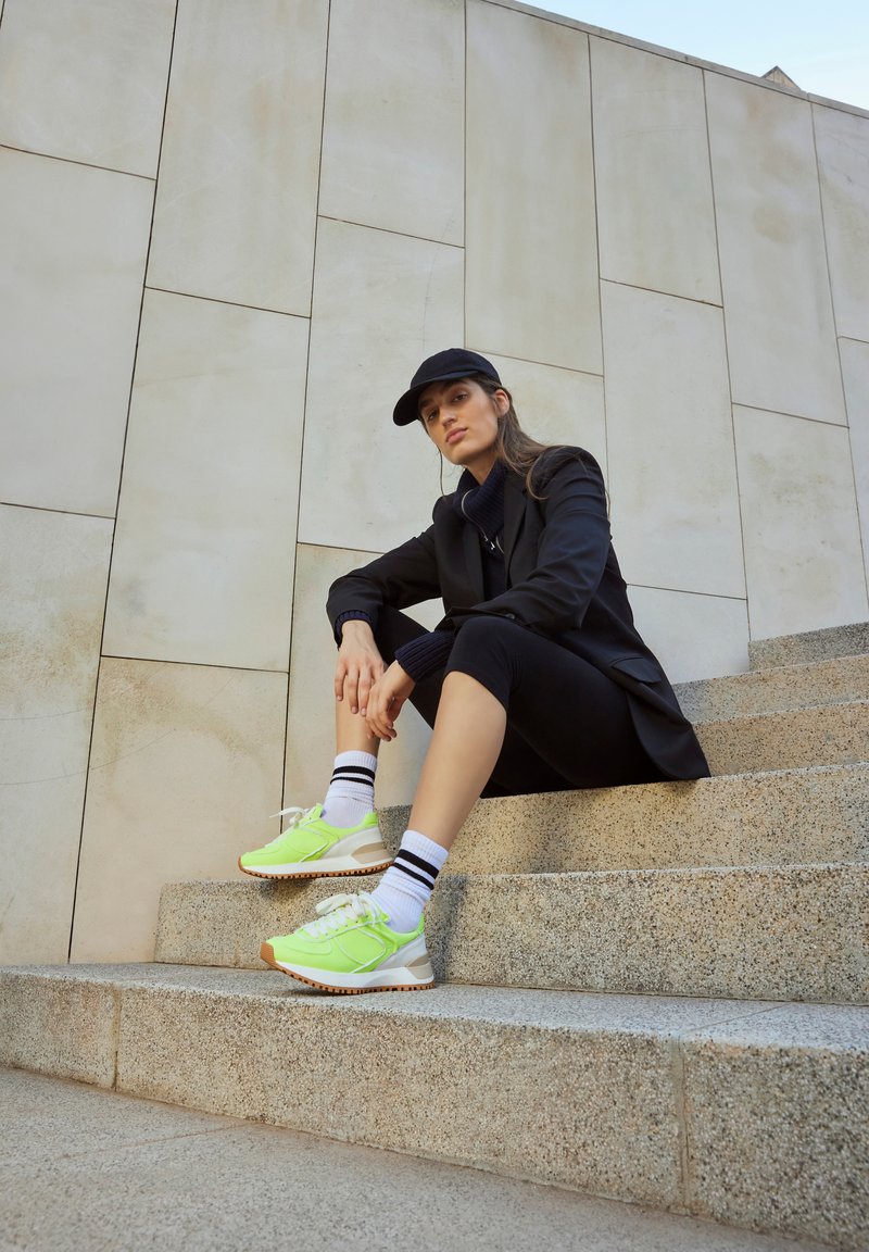 Zapatillas deportivas verde claro con cordones blancos y suelas marrones, combinadas con calcetines blancos con rayas negras. Modelo sentado en escalones de piedra.