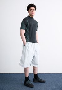 Camisa deportiva de manga corta de color verde oscuro con cuello alto, combinada con pantalones cortos de un gris claro. Zapatillas negras y calcetines oscuros completan el atuendo.
