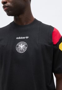 Homme portant un t-shirt noir Adidas avec le logo du Deutscher Fußball-Bund et des bandes rouges, noires et jaunes sur la manche.