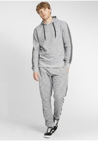 Sivi hoodie i odgovarajuće trenirke izrađeni od mekog materijala, sa crnim prugama duž strana i crnim vezicama. Crne tenisice upotpunjuju izgled.