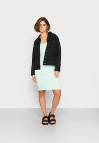 Jaqueta de denim preta sobre um vestido verde menta com texturas verticais. A modelo usa sandálias pretas com detalhes em fivela. Formas suaves e ajustadas com cores contrastantes.