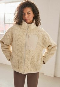 Cremfarbener Fleece-Jacke mit hohem Kragen, durchgehendem Reißverschluss und strukturiertem Finish. Verfügt über eine cremefarbene Stofftasche auf der linken Brust.
