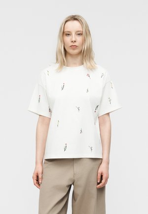 Ung kvinde med glat blondt hår iført hvid kortærmet skjorte med små blomsterprint og beige bukser, stående mod en ensfarvet baggrund.