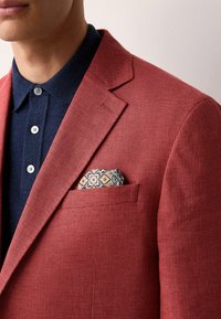 Rot strukturierter Blazer mit Spitzrevers, kombiniert mit einem marineblauen Hemd mit Knöpfen. Das Einstecktuch zeigt ein komplexes geometrisches Muster in Blau, Orange und Creme.