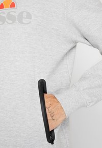 Sudadera gris con un logo y un bolsillo con cremallera negra en el costado. La tela parece suave con un acabado texturizado.