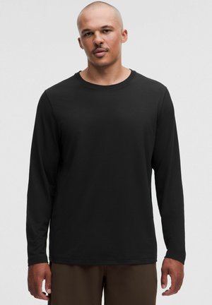 Mann mit rasiertem Kopf, der ein schlichtes schwarzes Langarmshirt und braune Hosen trägt, steht vor einem hellgrauen Hintergrund.