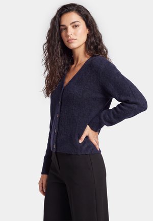 Femme aux longs cheveux bouclés porte un cardigan bleu marine boutonné et un pantalon noir, debout avec une main sur la hanche sur fond blanc.