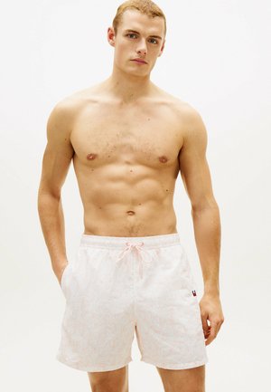 Skjorteløs mand med kort hår iført lyserøde badeshorts med mønster og sidelommer, stående foran en ensartet hvid baggrund.