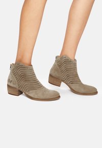 Bottes courtes en cuir taupe avec design perforé, bout arrondi et petit talon empilé. Dotées d'une fermeture éclair et d'un ornement papillon décoratif.