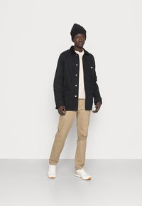 Chaqueta de pana negra con cuello, suéter beige, pantalones caqui y zapatillas blancas con acentos naranjas. Lleva puesto un gorro de punto negro.