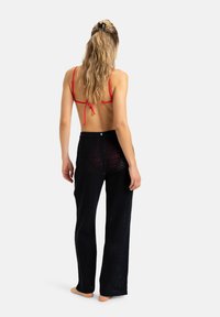Zwarte, brede broek met een gestructureerd, zigzagpatroon; gecombineerd met een rode top met bandjes op de rug. Model met de rug naar de camera, blootsvoets, haar gestyled.
