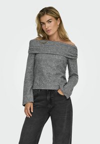 Grauer schulterfreier Pullover aus weich stricktem Material, mit langen Ärmeln und tailliertem Oberteil. Kombiniert mit hochgeschnittenen schwarzen Jeans.