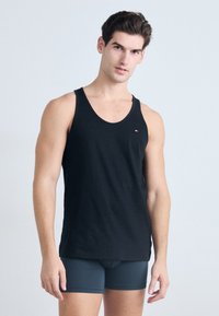Svart tanktopp laget av bomull, med rund halslinning og en liten logoaccent. Modellen har på seg grå tettsittende shorts.