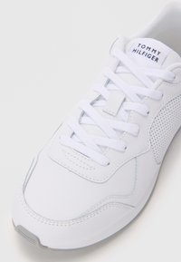 Fehér sneaker sima bőr felsőrésszel, perforált díszítésekkel, lapos fűzőkkel, párnázott nyelvvel és szürke gumitalppal.