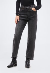 Άτομο που φοράει ξεθωριασμένο μαύρο τζιν ψηλόμεσο με πλευρικές σκισμές στο τελείωμα, μαύρο εφαρμοστό μακρυμάνικο τοπ, μαύρες μποτάκια με τακούνι και βραχιόλι.