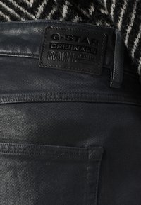 Pantalones vaqueros de mezclilla negra con una textura suave, que presentan una etiqueta de cuero G-Star Originals en la cintura y un bolsillo trasero.