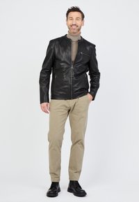 Man die glimlacht, in een zwart leren jack, een beige coltrui, kaki broeken en zwarte laarzen, staand tegen een eenvoudige lichte achtergrond.