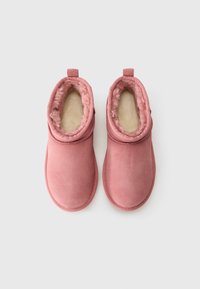 Une paire de chaussons en daim rose avec des intérieurs doublés de laine douce, montrés de dessus sur un fond blanc.