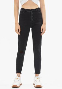 Schwarze hochgeschnittene Skinny-Jeans mit einem Knopfverschluss vorne, leichtem Riss am Knie und fransigem Saum, kombiniert mit weißen Sneakers.