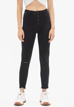 Jeans skinny noirs taille haute avec une fermeture à bouton sur le devant, une légère déchirure au genou et un ourlet effiloché, associés à des baskets blanches.