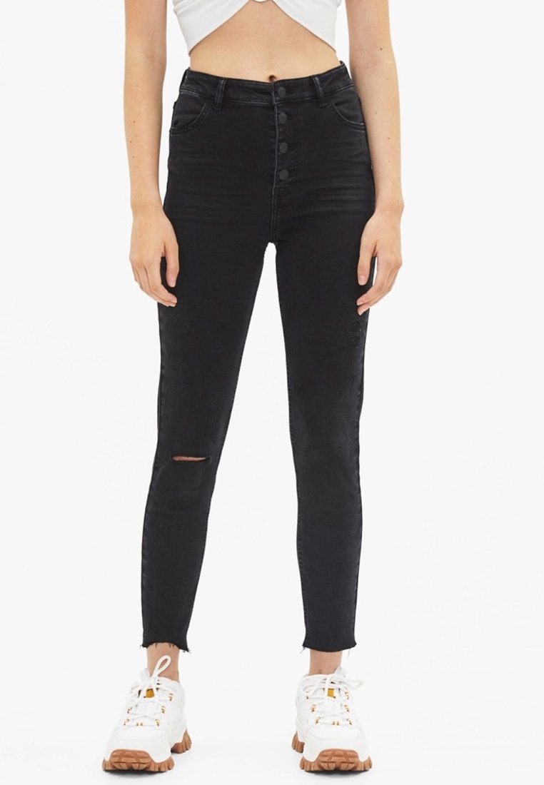 Schwarze hochgeschnittene Skinny-Jeans mit einem Knopfverschluss vorne, leichtem Riss am Knie und fransigem Saum, kombiniert mit weißen Sneakers.