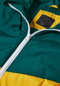 Bunte Jacke mit einem türkisfarbenen oberen Bereich und einem gelben unteren Bereich, mit einem weißen Reißverschluss und einem schwarzen Etikett innen. Glatte Textur.